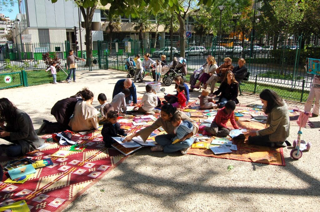 Bibliothèque de rue plan large, de nombreux enfants et adultes sur des tapis colorés dans un square au soleil, avec de nombreux livres au sol, des adultes assis sur un banc et des poussettes à l'arrière plan.