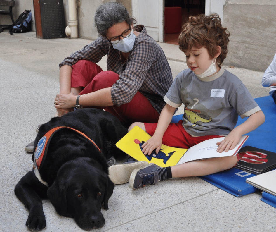 Enfant assis en situation de lecture avec un adulte et un chien. L'enfant a les mains posées sur les pages tactiles (une sur le texte l'autre sur l'illustration) L'adulte regarde l'enfant.